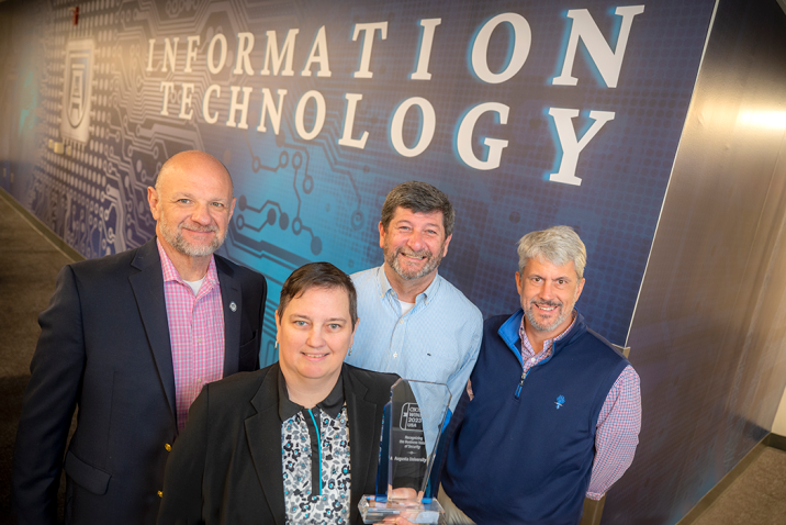Four people stand in front of a wall with the words "Information Technology" while holding an award.