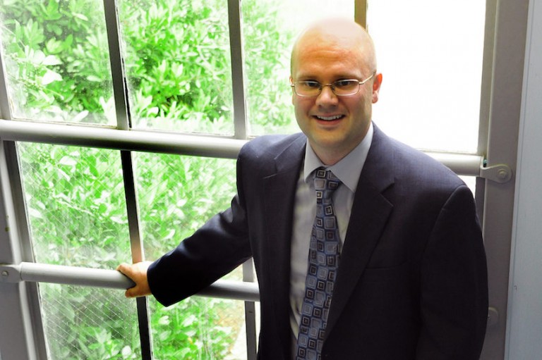 Dr. Brian Miller, wearing a blue suit and glasses, stands in front of window