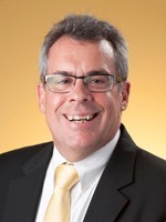 A man in a white shirt, gold tie and black suit coat wearing glasses smiles at the camera