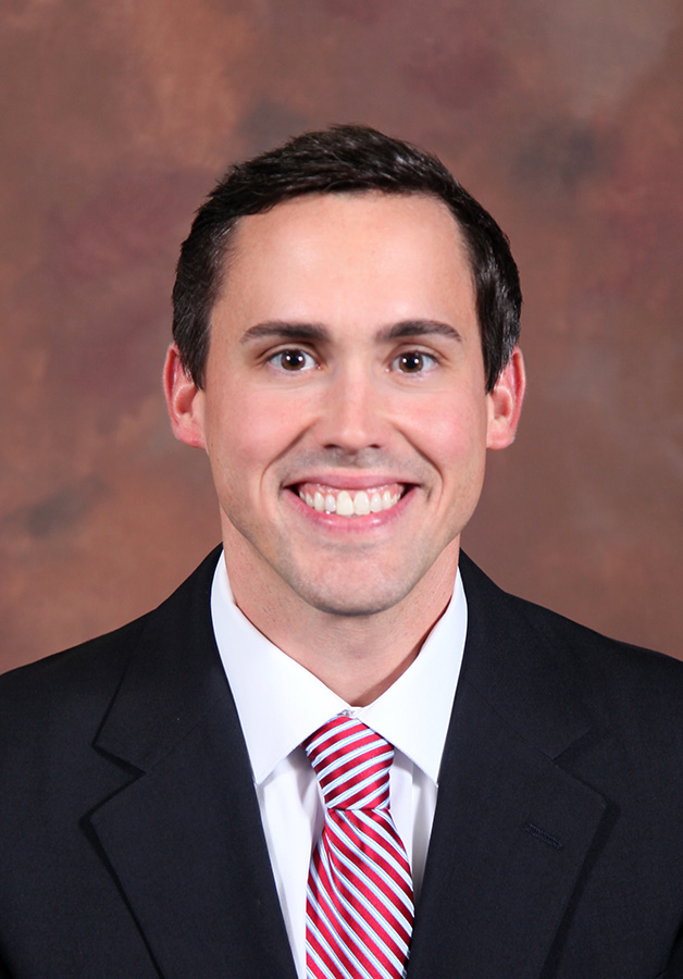 A man in a white shirt, red and white striped tie and black suit coat smiles at the camera