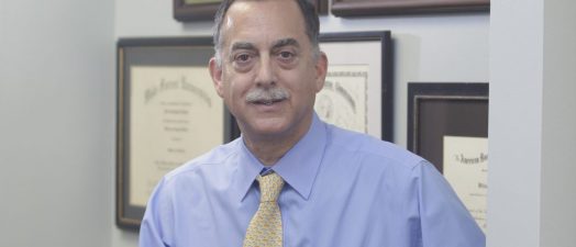 Dr. Vaughn McCall, wearing a blue shirt and yellow tie, sits on the corner of a desk in his office