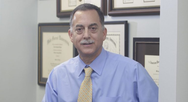 Dr. Vaughn McCall, wearing a blue shirt and yellow tie, sits on the corner of a desk in his office