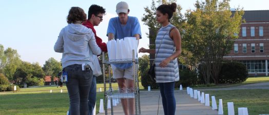 Four volunteers standing over a cart of paper luminary bags. The sidewalk they're standing on is lit on each side by the luminary bags they placed.