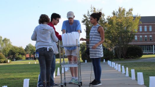 Four volunteers standing over a cart of paper luminary bags. The sidewalk they're standing on is lit on each side by the luminary bags they placed.