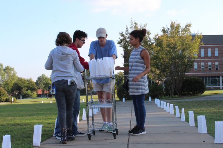 Four volunteers standing over a cart of paper luminary bags. The sidewalk they're standing on is lit on each side by the luminary bags they placed.