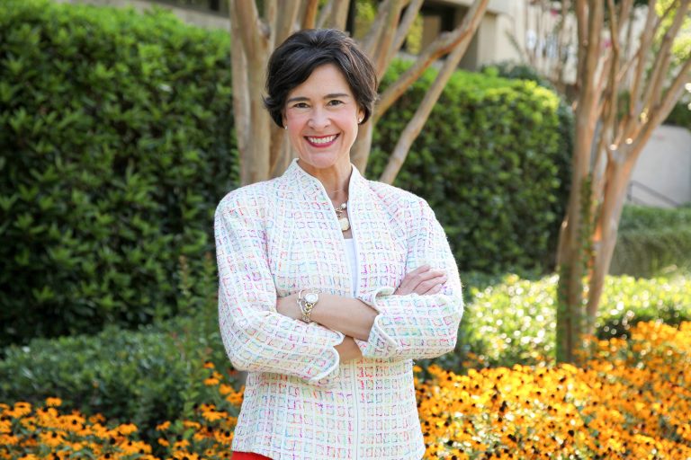 Color photo of woman standing in front of trees and orange flowers