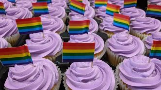 Photo of vanilla cupcakes with purple frosting. Cupcakes are decorated with mini LGBTQ+ pride flag on toothpicks.