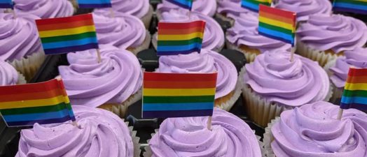 Photo of vanilla cupcakes with purple frosting. Cupcakes are decorated with mini LGBTQ+ pride flag on toothpicks.