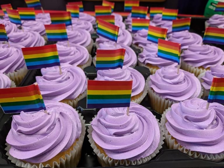 Photo of vanilla cupcakes with purple frosting. Cupcakes are decorated with mini LGBTQ+ pride flag on toothpicks.