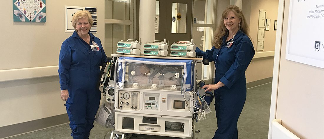 Two transport nurses dressed in blue coveralls roll a baby isolette on wheels