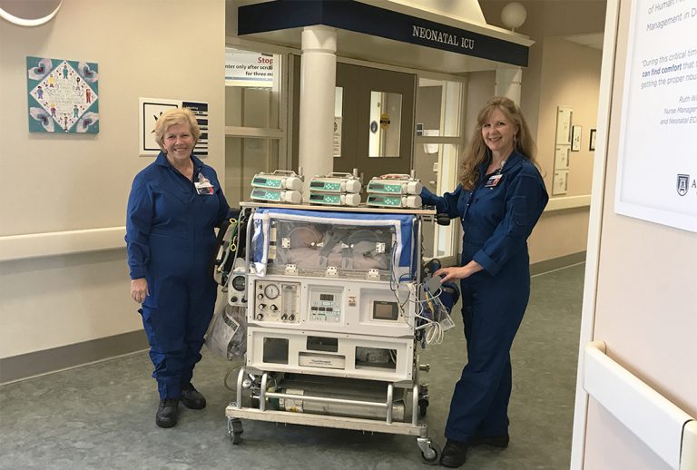 Two transport nurses dressed in blue coveralls roll a baby isolette on wheels