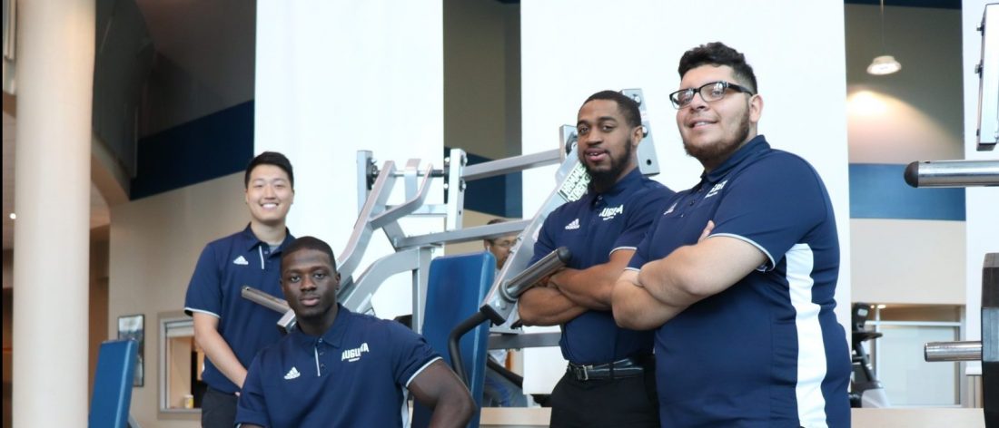Four male students in matching blue polos, posing in front of fitness equipment