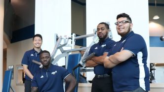 Four male students in matching blue polos, posing in front of fitness equipment