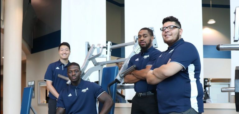 Four male students in matching blue polos, posing in front of fitness equipment