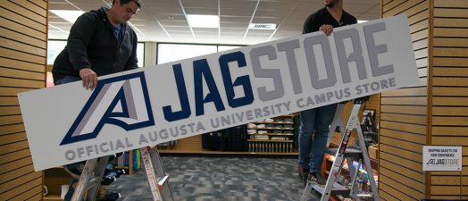 Two men hang a sign identifying the Augusta University JagStore.