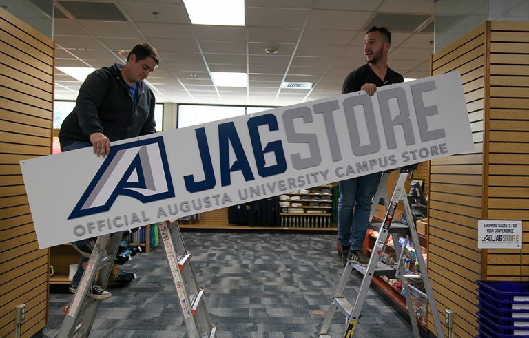 Two men hang a sign identifying the Augusta University JagStore.