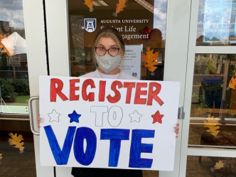 image of a woman in a grey mask holding a white sign that reads "REGISTER TO VOTE," with stars on it