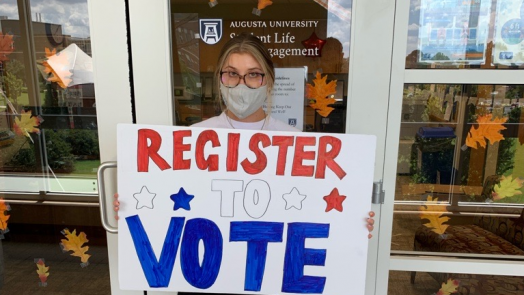 image of a woman in a grey mask holding a white sign that reads "REGISTER TO VOTE," with stars on it