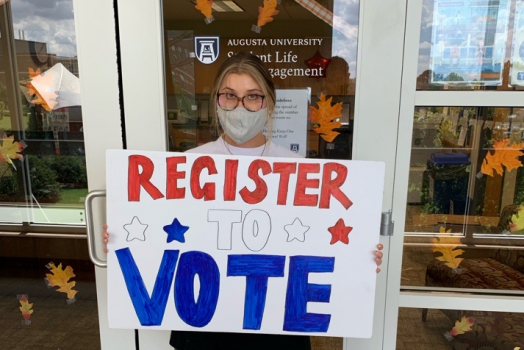 image of a woman in a grey mask holding a white sign that reads "REGISTER TO VOTE," with stars on it
