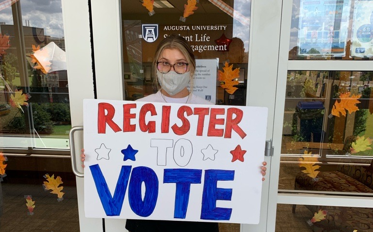 image of a woman in a grey mask holding a white sign that reads "REGISTER TO VOTE," with stars on it
