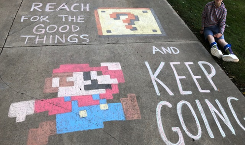 Student sits beside driveway chalk art. Art is a drawing of Mario, captioned "Reach for the good things and keep going."