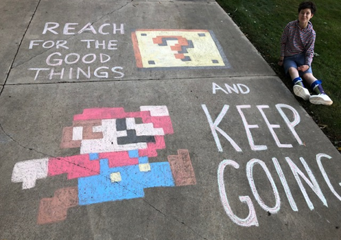 Student sits beside driveway chalk art. Art is a drawing of Mario, captioned "Reach for the good things and keep going."