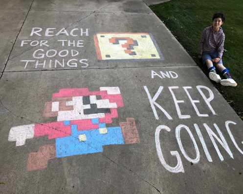 Student sits beside driveway chalk art. Art is a drawing of Mario, captioned "Reach for the good things and keep going."