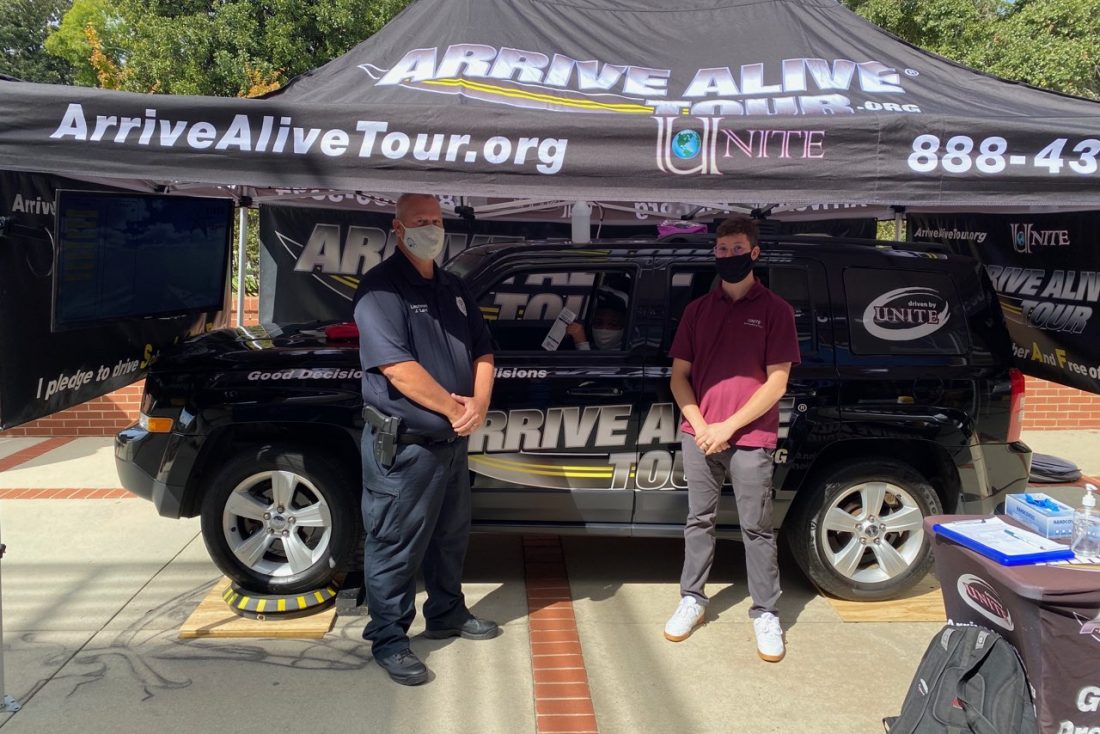 wearing masks, a male police officer and man stand under a tent, in front of a car with "Arrive Alive Tour" branding on it