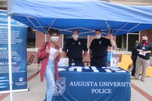 all wearing masks, a female student and two male police officers smile in front of an "Augusta University Police" tent