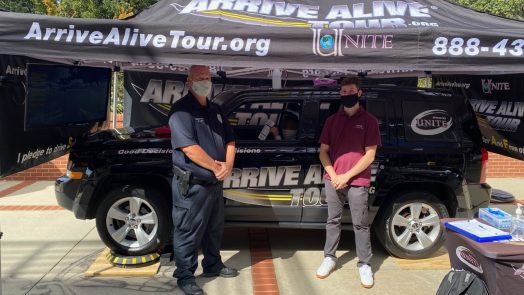 wearing masks, a male police officer and man stand under a tent, in front of a car with "Arrive Alive Tour" branding on it