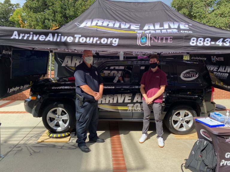 wearing masks, a male police officer and man stand under a tent, in front of a car with "Arrive Alive Tour" branding on it