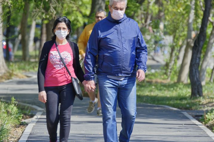 Older man and woman holding hands while walking and wearing face masks.