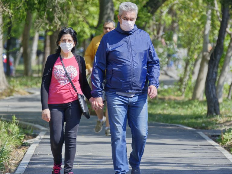 Older man and woman holding hands while walking and wearing face masks.