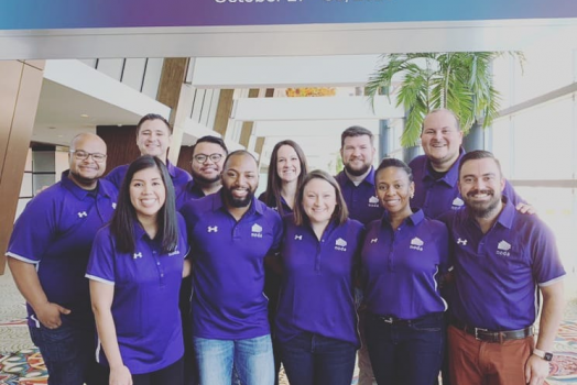 11 men and women in purple collared shirts smile for a photo