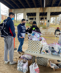 three people wearing facemasks stand beside a table filled with bags of supplies