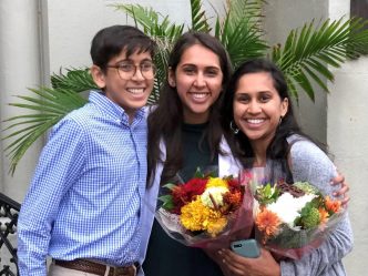 A brother and his two sisters, one sister holding flowers, smile for a photo