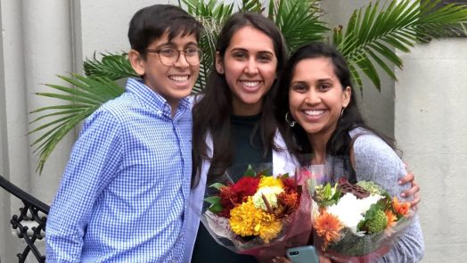 A brother and his two sisters, one sister holding flowers, smile for a photo