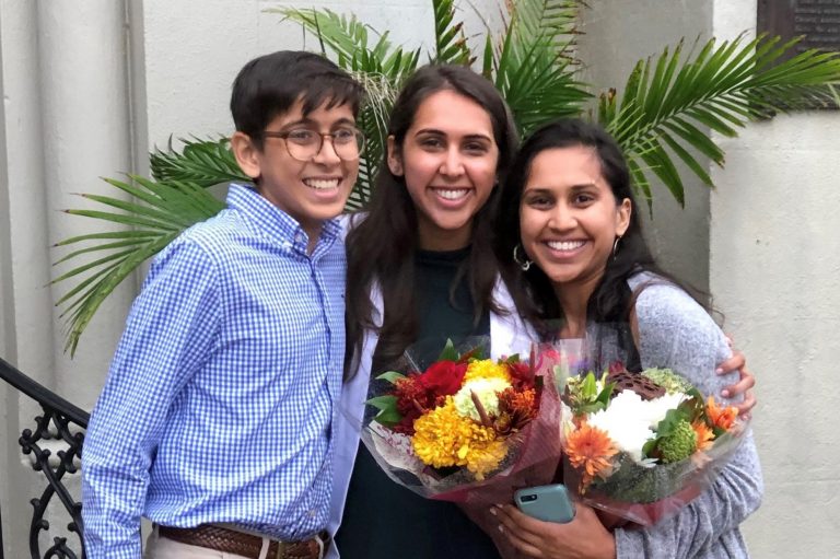 A brother and his two sisters, one sister holding flowers, smile for a photo