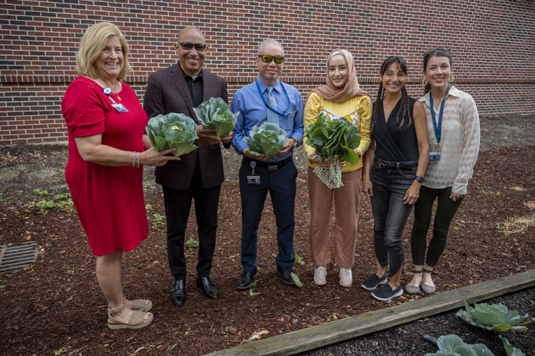 6 people stand in mulch in a vegetable garden, holding leafy greens