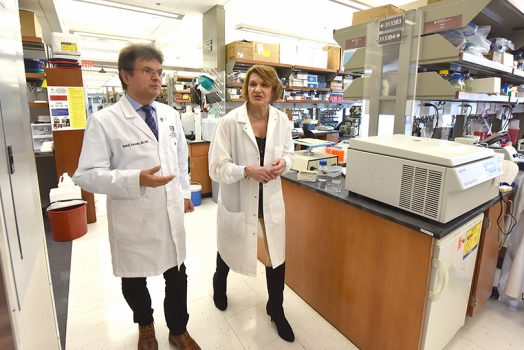 Two doctors (male on the left and female on the right) wearing white coats walk together through a lab