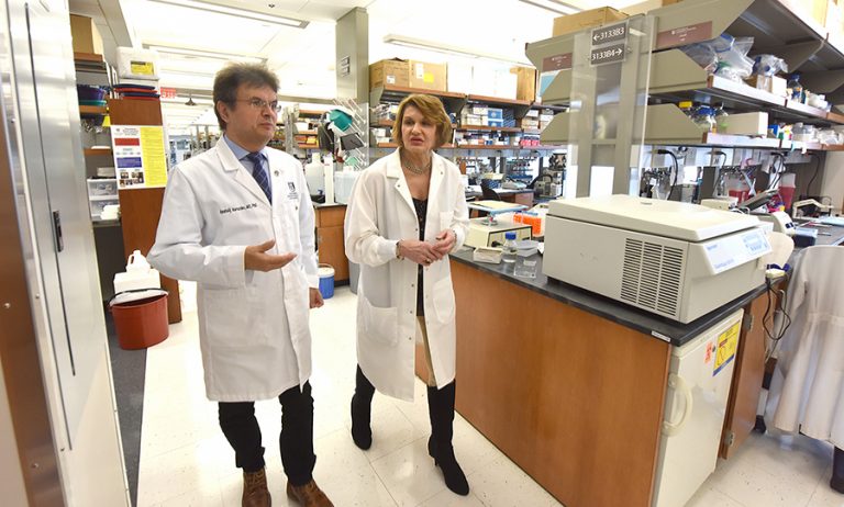 Two doctors (male on the left and female on the right) wearing white coats walk together through a lab