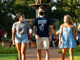two women and one man, all wearing face masks, walking outside near a fountain