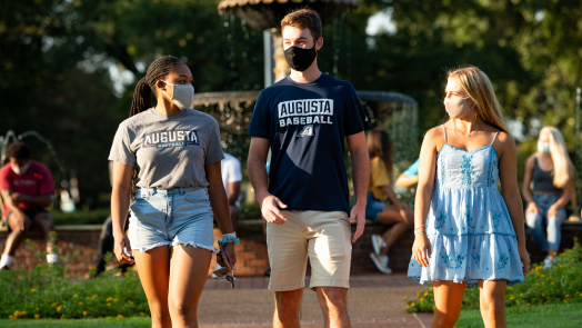 two women and one man, all wearing face masks, walking outside near a fountain