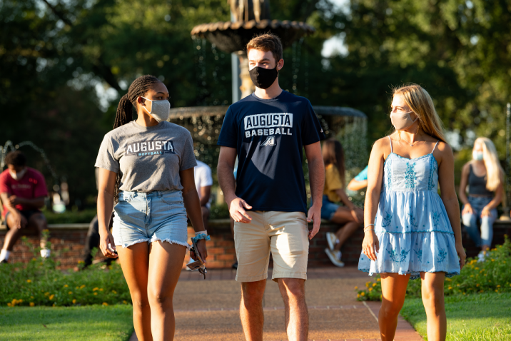 two women and one man, all wearing face masks, walking outside near a fountain