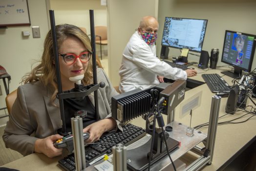 Woman with red glasses rests chin on machine used to break camouflage while a physician in a white coat looks on in the background