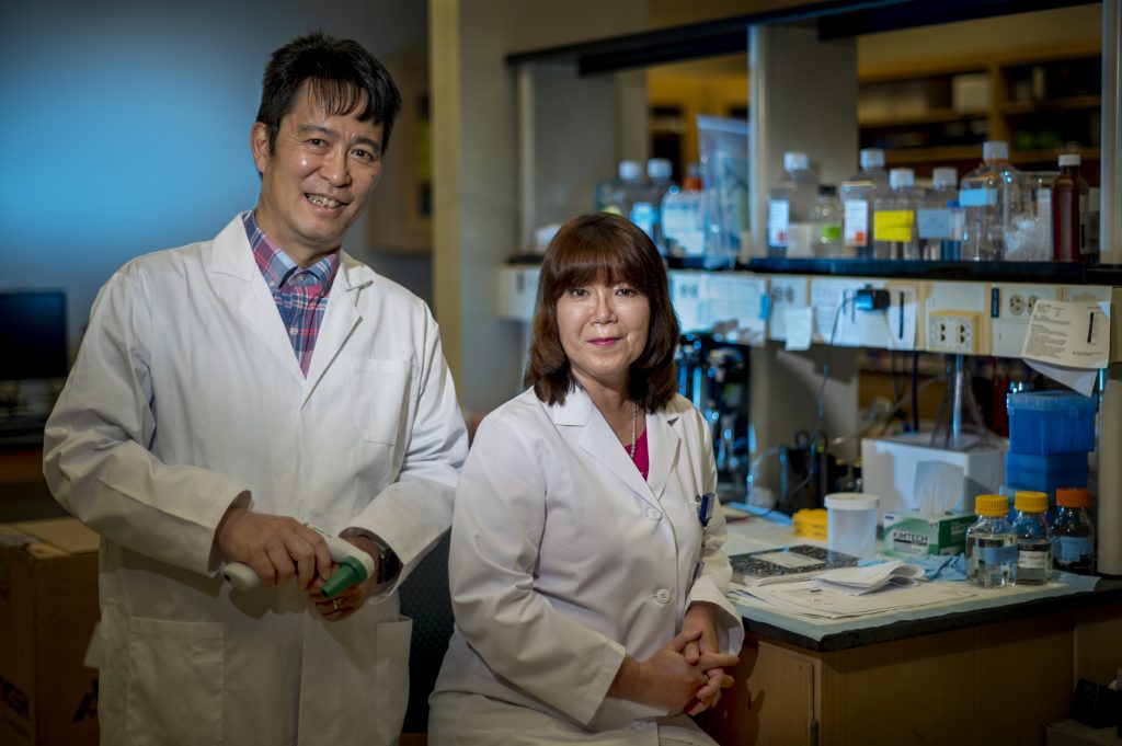 Two researchers, a man on the left and woman on the right, stand in a lab with white coats on.