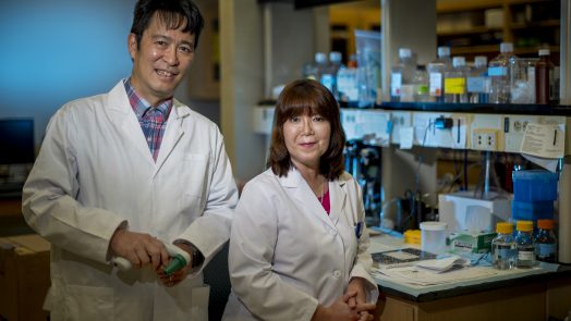 Two researchers, a man on the left and woman on the right, stand in a lab with white coats on