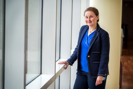 Woman in blue suit with blue blouse stands in front of windows looking at camera