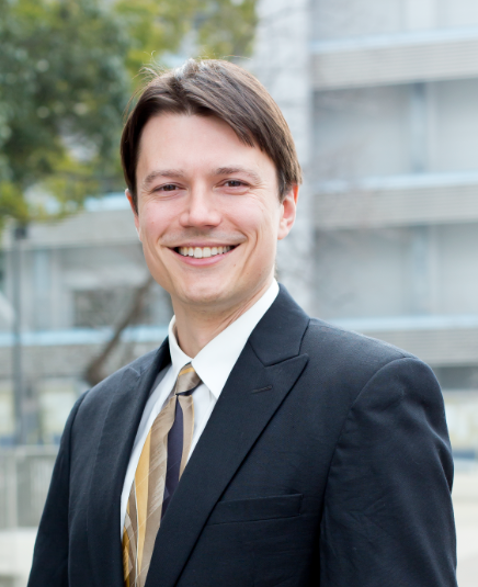 Man standing in black suit standing in front of building smiling.