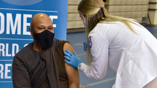 woman in white coat giving a shot to person sitting in a chair, masked